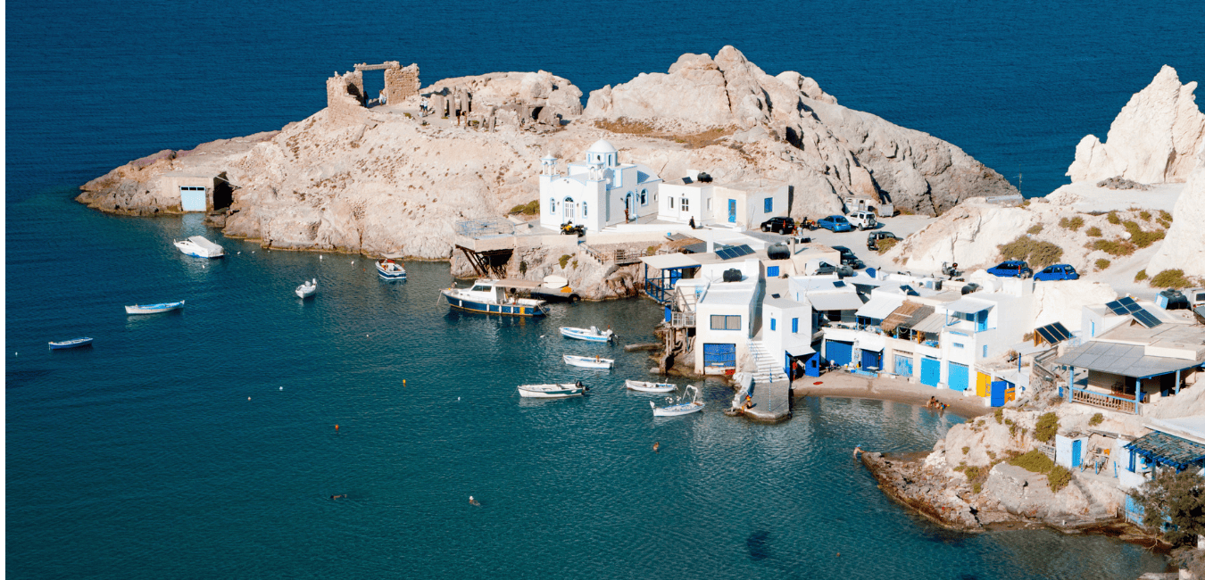 Boats anchored in the shallows of the coast of Milos with white houses on the coast